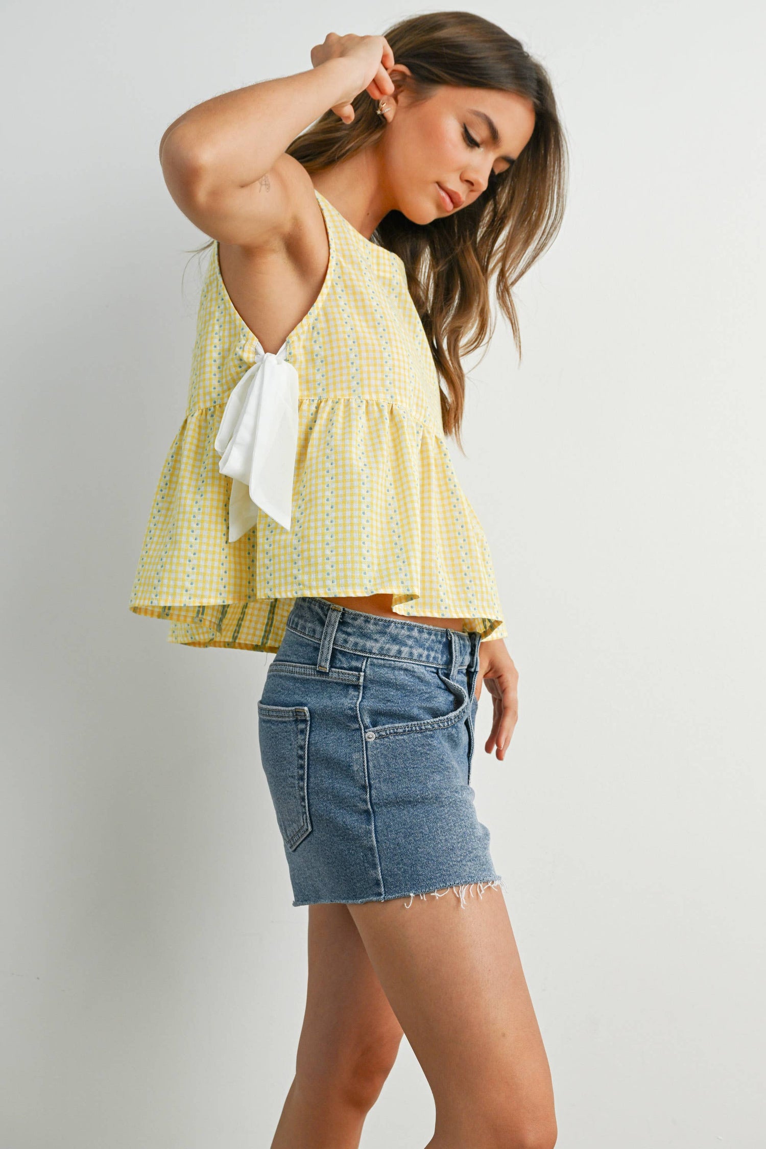 girl with yellow top with white ties and jean shorts holding hair behind her ear as a cover picture for the tops
